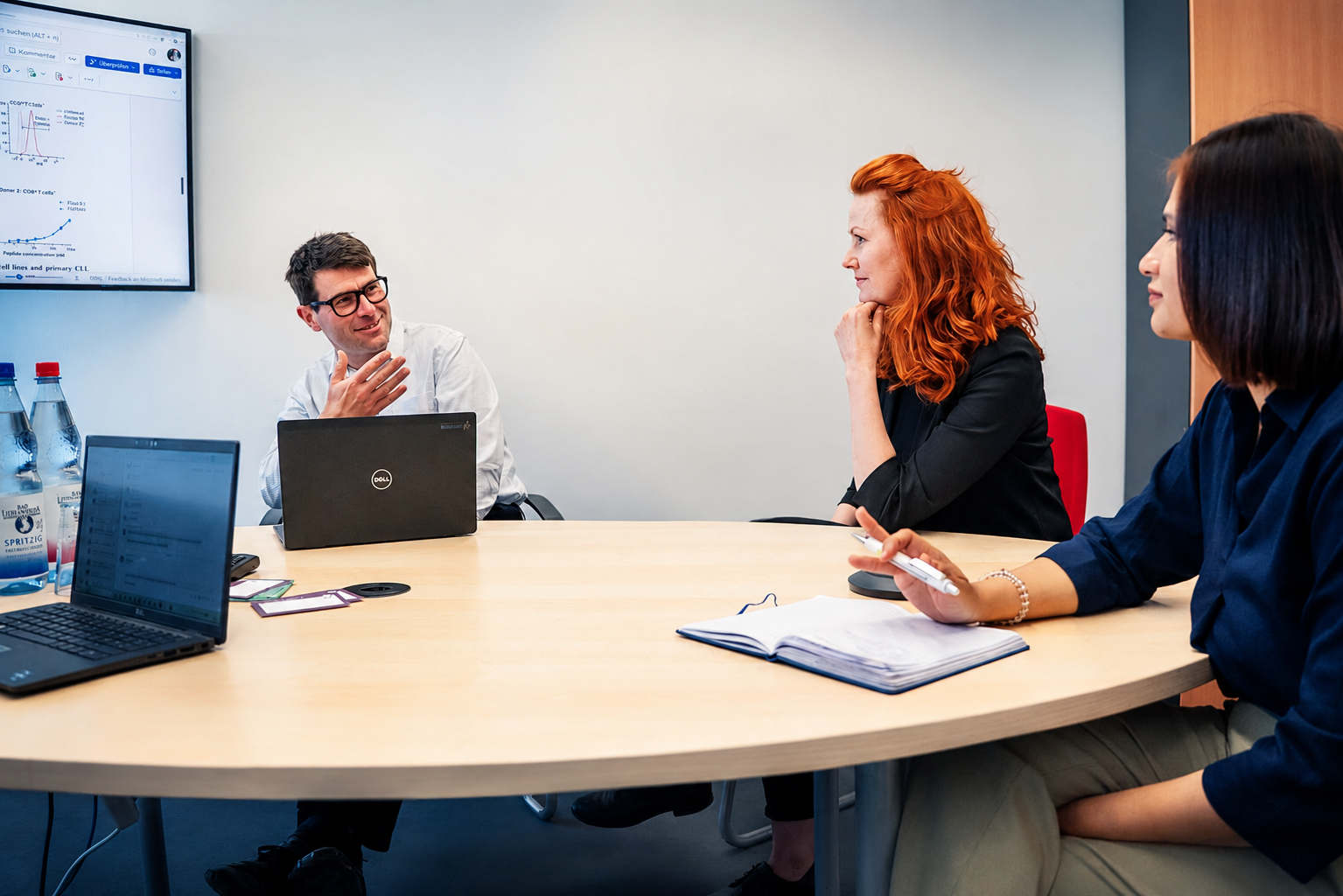Three people sit at a conference table in a meeting room, discussing in front of a screen showing charts.
